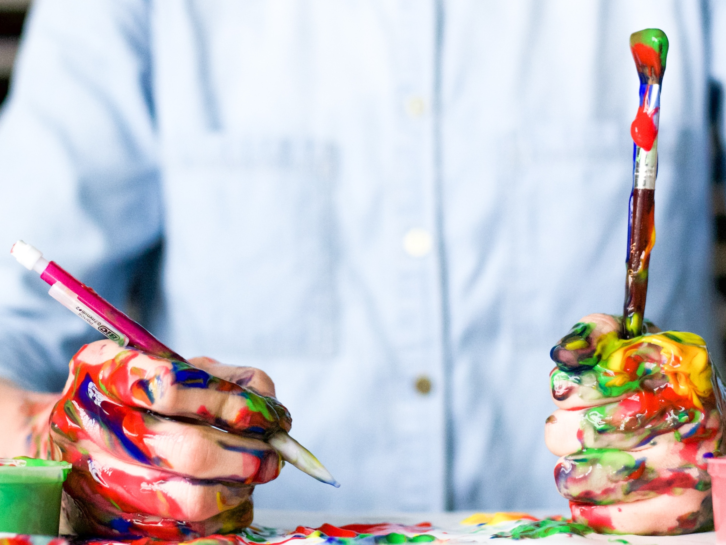 A person holding a paintbrush in their left hand fist and a pen in their right with both hands covered in red, green , blue, yellow splatters of thick paint with three paint pots in the foreground
