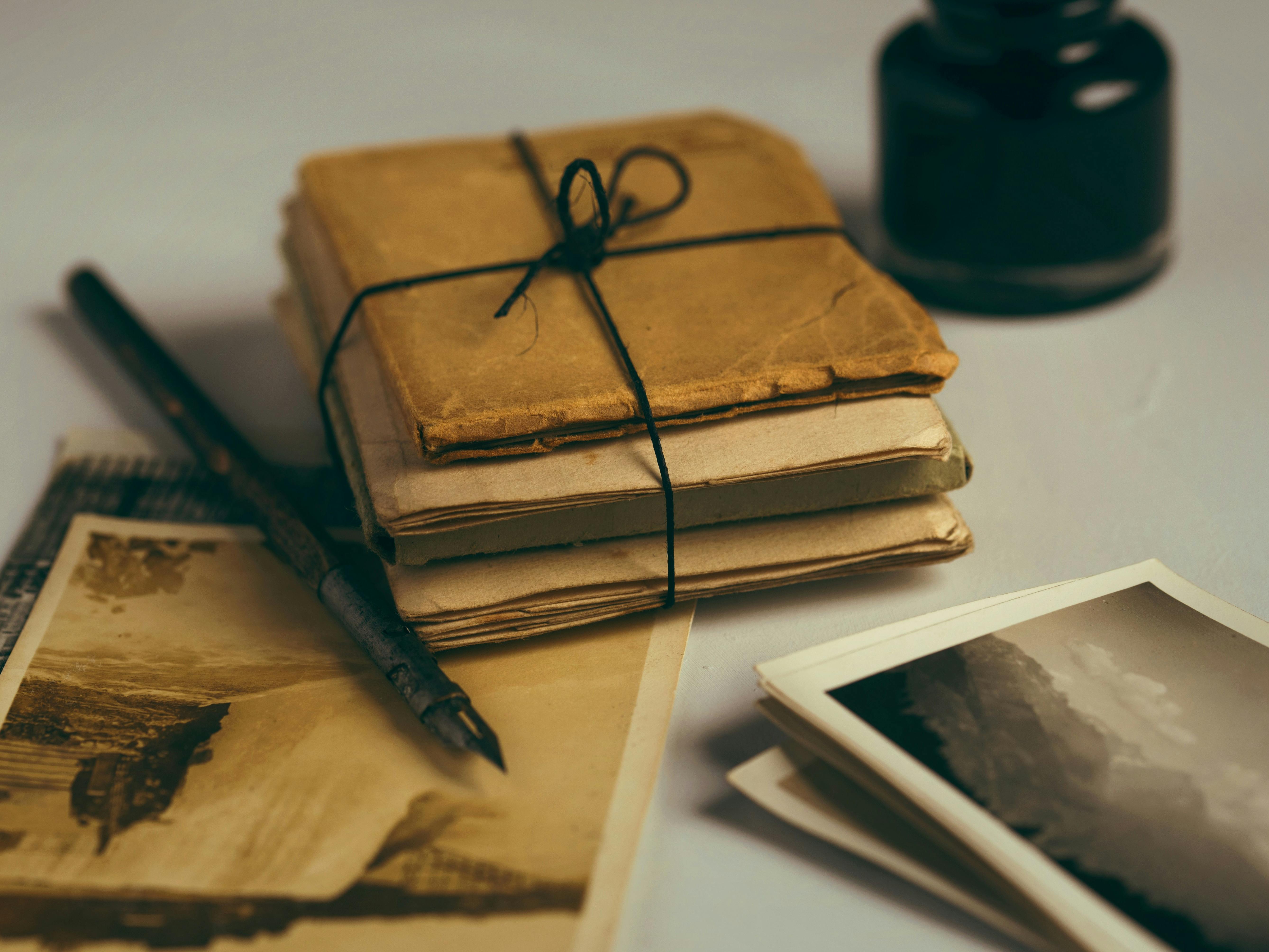An image of a collection of aged items, these include a laced stack of documents and aged brown photographs, a fountain pen and pot of black ink in the background