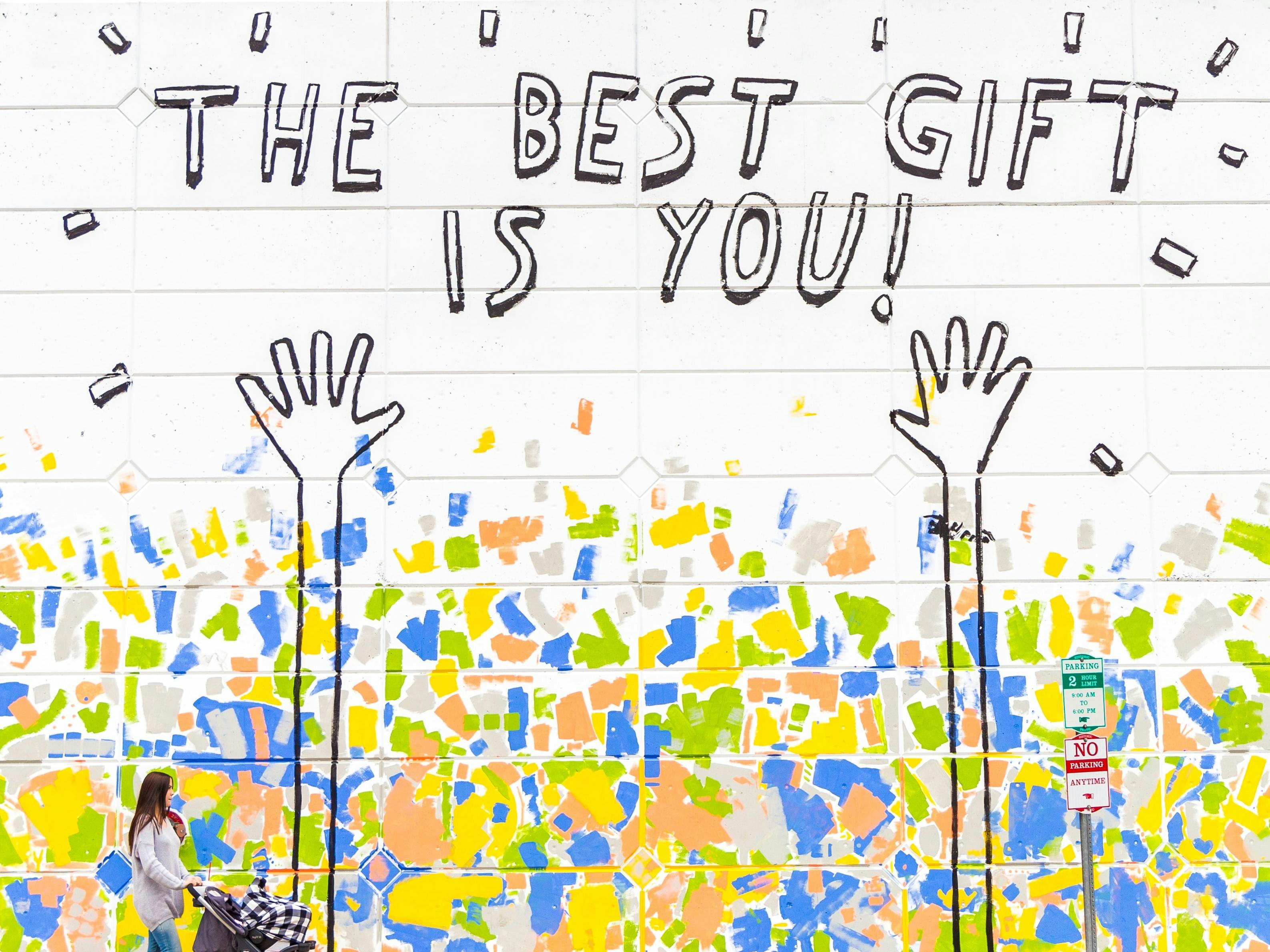 Mural of two upreached arms and the words 'The Best Gift is You' with green, pink, yelllow and blue splashes on a wall and a woman with pram walking from the left of the picture towards the right