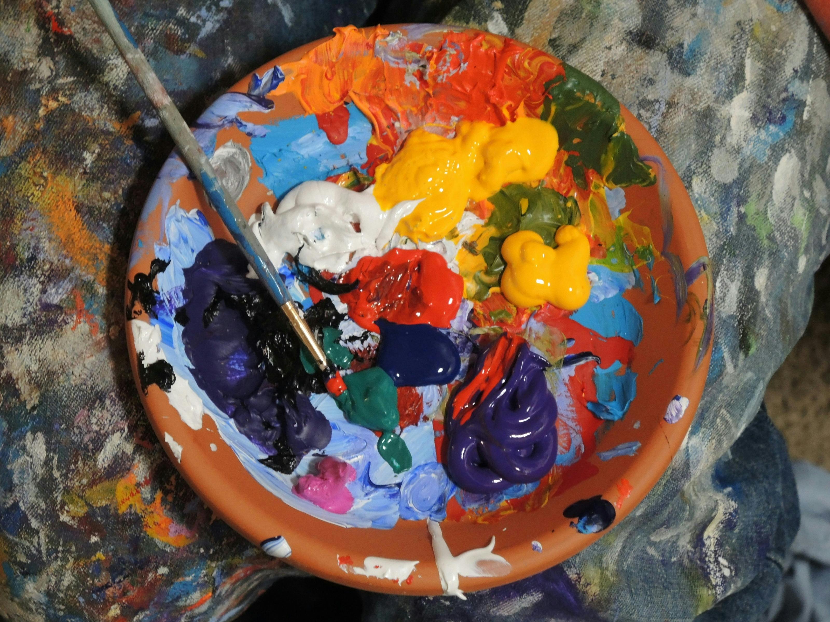 A dish filled with different coloured pain including yellow, white, red, blue and green with a used paint brush with paint splattered fabric