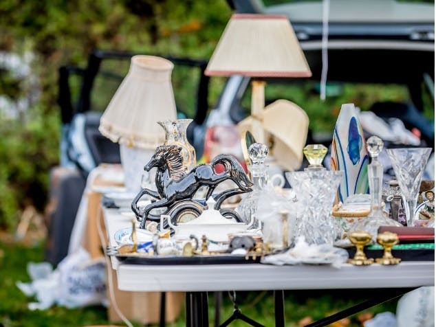Photograph of a table filled with vases, lamps and ornaments set out in a car boot style with an open car boot in the background