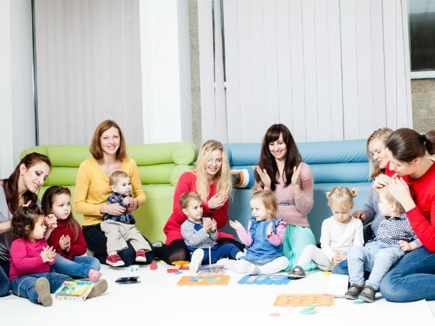 Photograph of 6 women with 7 toddlers and preschoolers sat in a half circle with children and women clapping hands like attending a rhymme and sing club