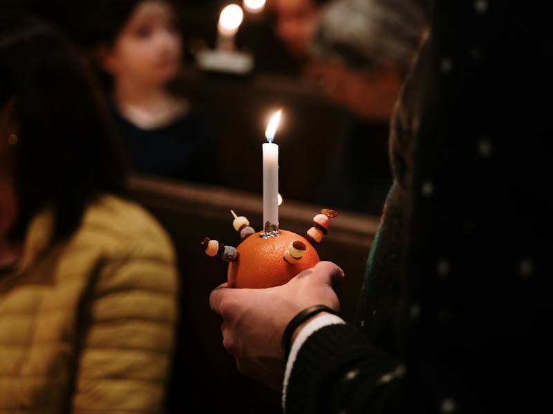Image courtesy of Al Elmes on Unsplash Photograph of a person holding a Christingle candle - an orange with four cocktail sticks attached to the fruit with threaded sweets and raisons and a lit candle in the centre