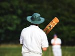 Cricket player with green cricket hat and bat with their back to the camera and a individual in cricket whites in the background