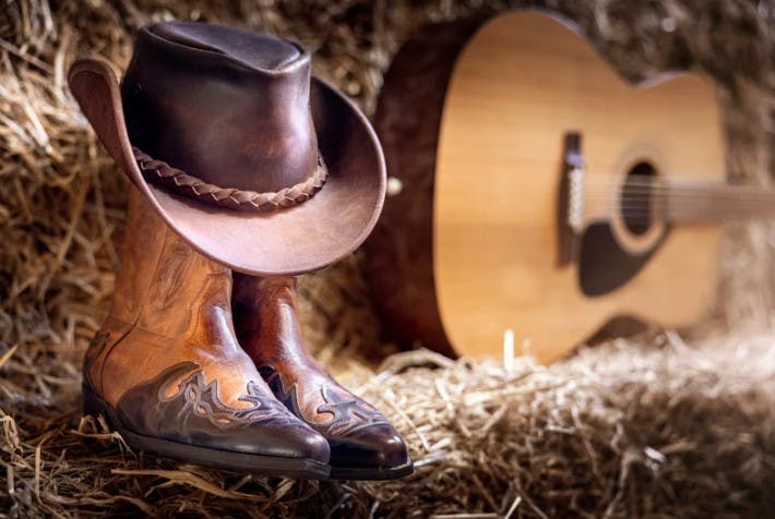 Photograph of a cowboy hat, boots and a guitar on straw bales