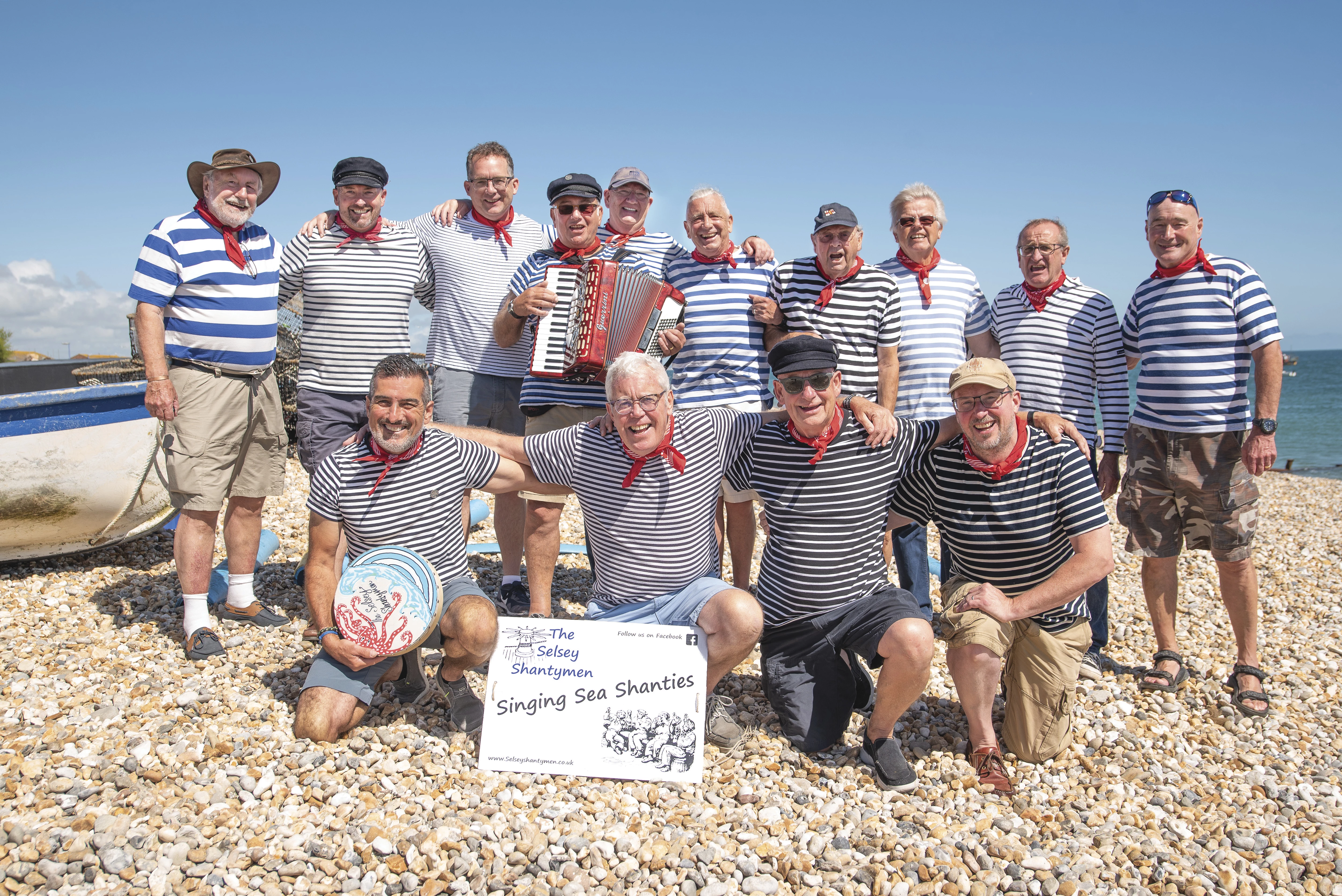 A picture of 14 local men from Selsey on the beach all dress in blue and white striped tops with red handkerchiefs. One has an acordian and there is a sign at the front with their singing group name 'The Selsey Shantymen'