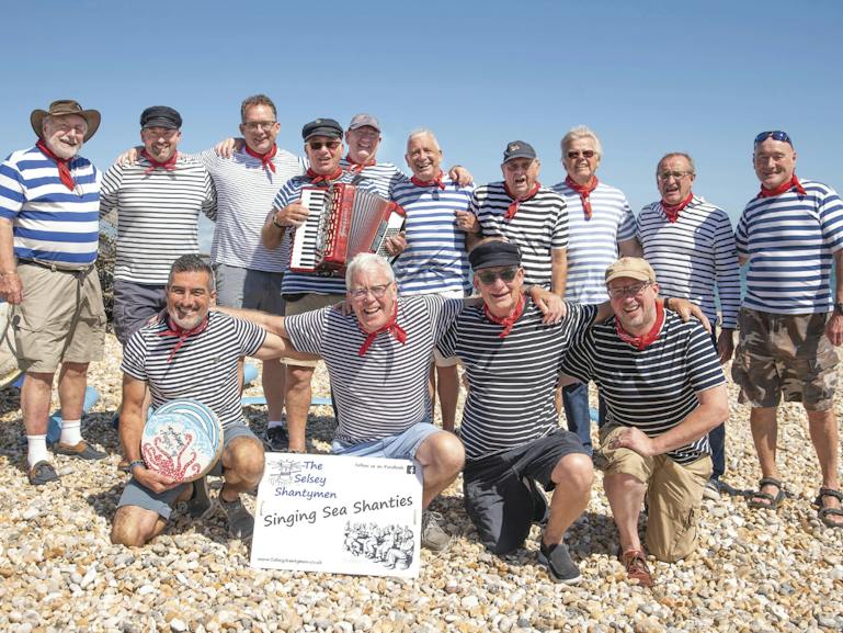 The Selsey Shantymen 2026 A picture of 14 local men from Selsey on the beach all dress in blue and white striped tops with red handkerchiefs. One has an acordian and there is a sign at the front with their singing group name 'The Selsey Shantymen'