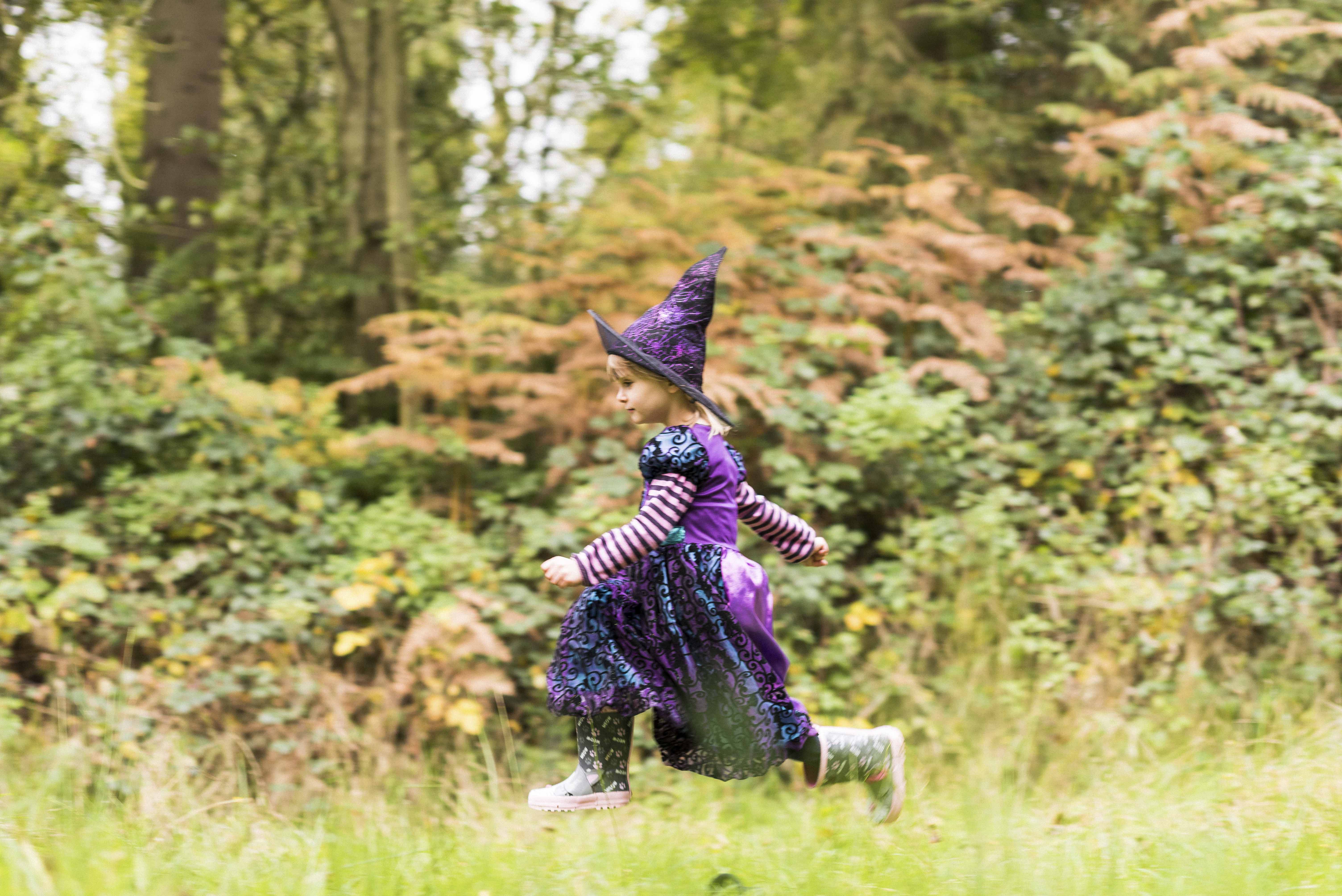 A young child in a purple witch costume and wellies running through a field with the autumnal woods behind.