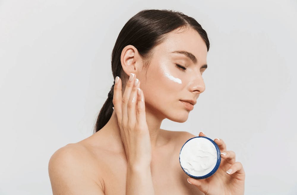 A woman applying a thick cream from a jar to her face, practicing a skin care routine for smooth and hydrated skin.