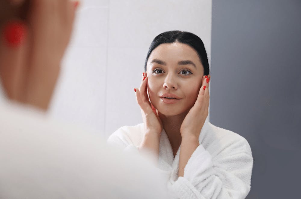 A woman in a white robe applying skin care product to her face while looking in the mirror, focusing on her skincare regimen.