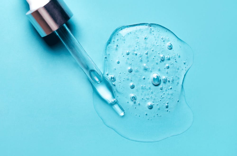 Close-up of a dropper dispensing hyaluronic acid serum on a blue surface, showcasing the product's watery texture and bubbles.