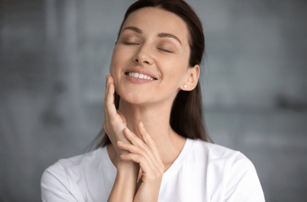 A smiling woman gently touching her face, with glowing skin, after applying a hyaluronic acid serum as part of her skincare routine.