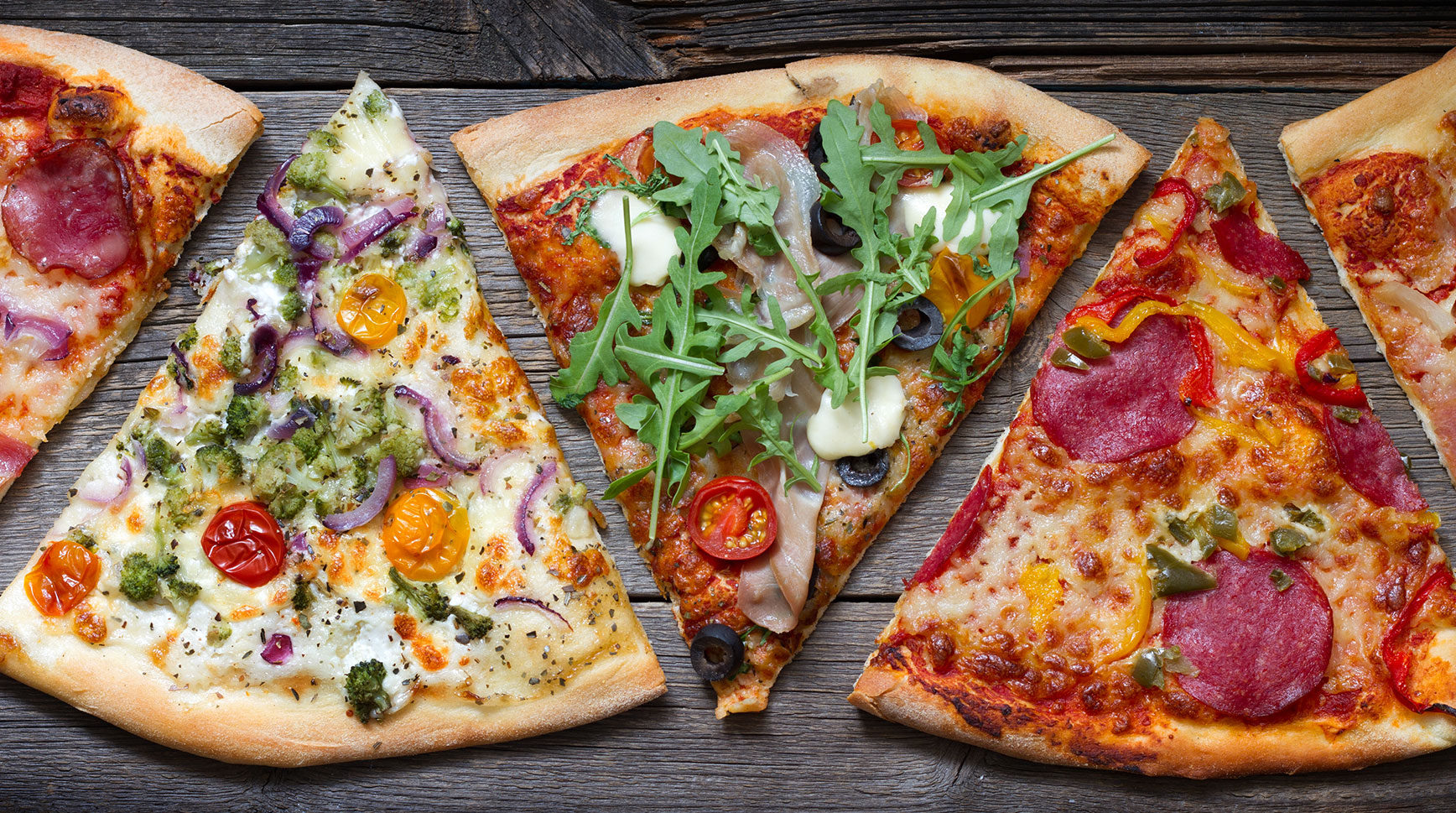 Pizza slices with different toppings, from prosciutto and arugula to salami and peppers, laid out on a wooden table