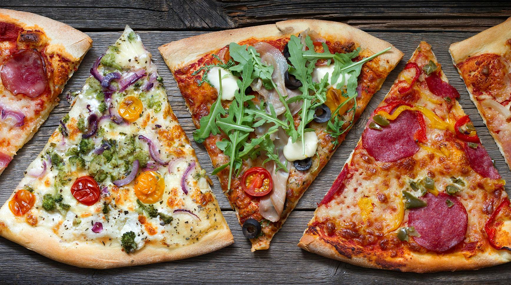 Pizza slices with different toppings, from prosciutto and arugula to salami and peppers, laid out on a wooden table