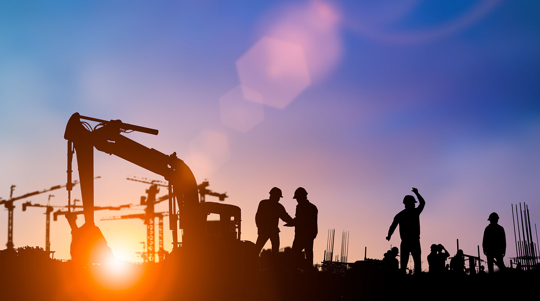 Silhouettes of construction workers and an excavator against a sunset sky with cranes in the background