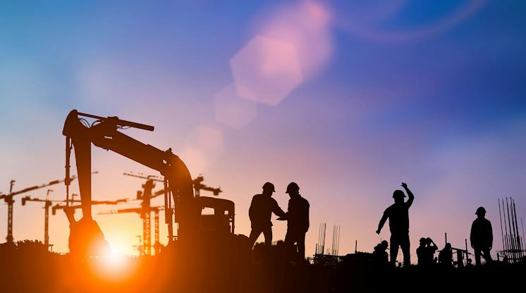 Silhouettes of construction workers and an excavator against a sunset sky with cranes in the background