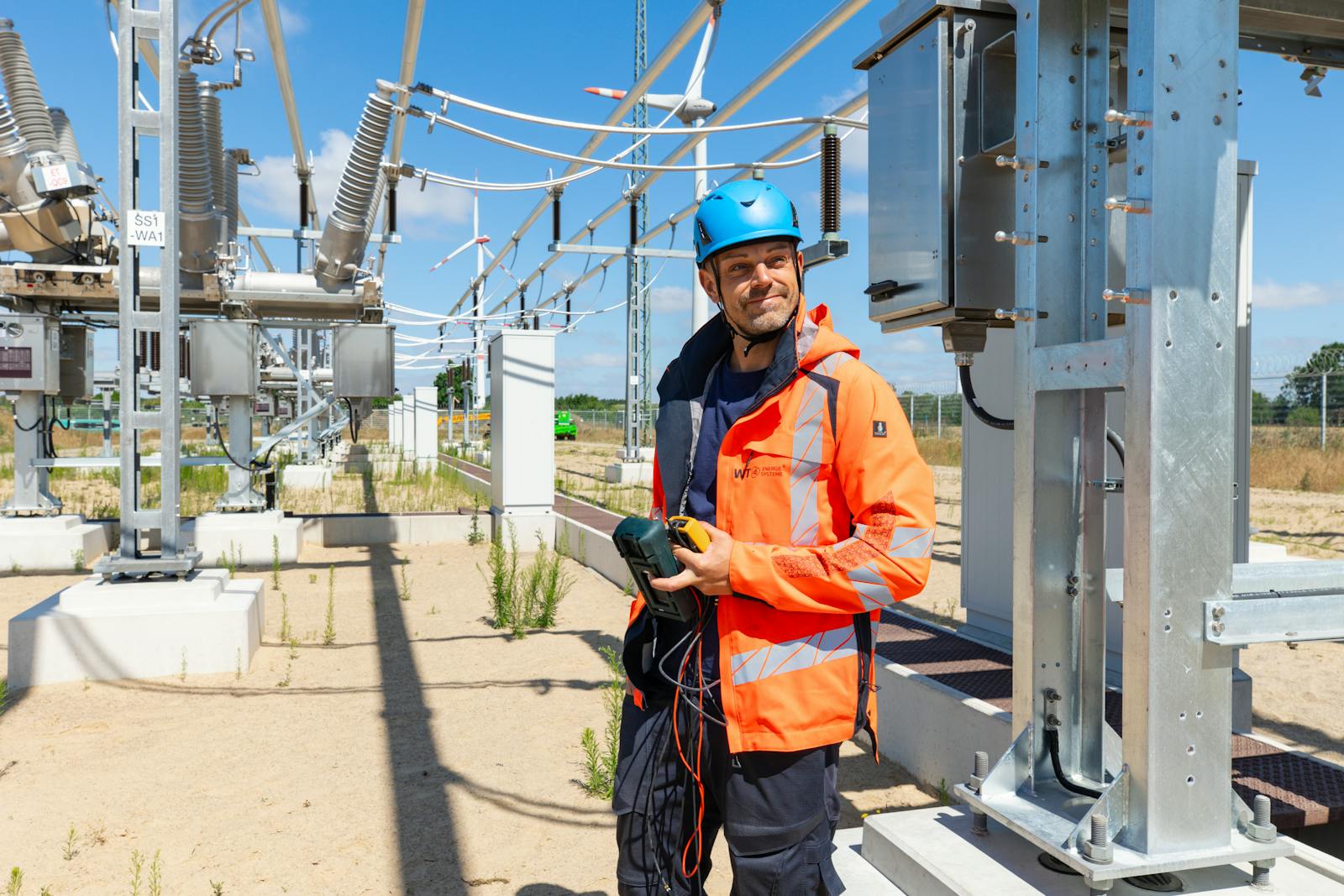 Mitarbeiter von WT Energiesysteme mit Messgerät bei der technischen Prüfung einer Hochspannungsschaltanlage im Umspannwerk, mit Windenergieanlagen im Hintergrund.