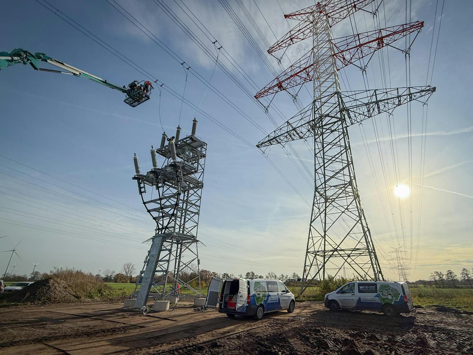WT Energiesysteme carrying out installation work on a high-voltage overhead line connection using an aerial work platform, with switchgear and a transmission tower.