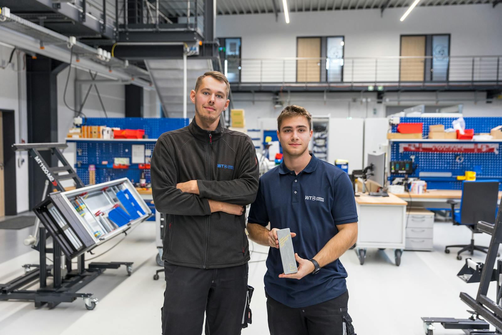 Zwei Mitarbeitende von WT Energiesysteme präsentieren in der Fertigungshalle die Auszeichnung des Unternehmens als „Most Responsible Employer“ durch kununu und DIE ZEIT.