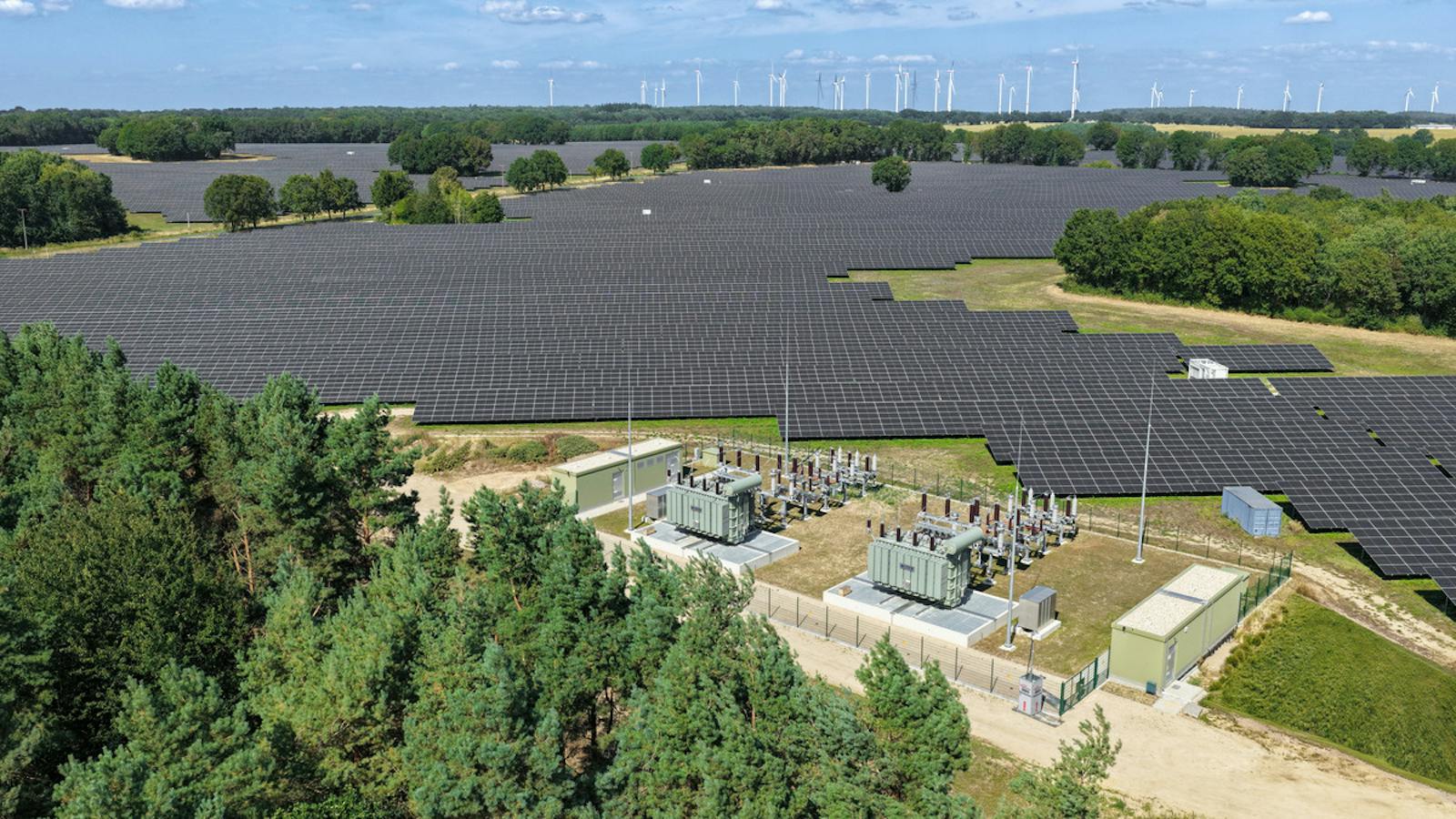 Luftaufnahme eines Umspannwerks mit zwei Leistungstransformatoren inmitten einer großflächigen Photovoltaikanlage. Im Hintergrund sind Windkraftanlagen zu sehen.