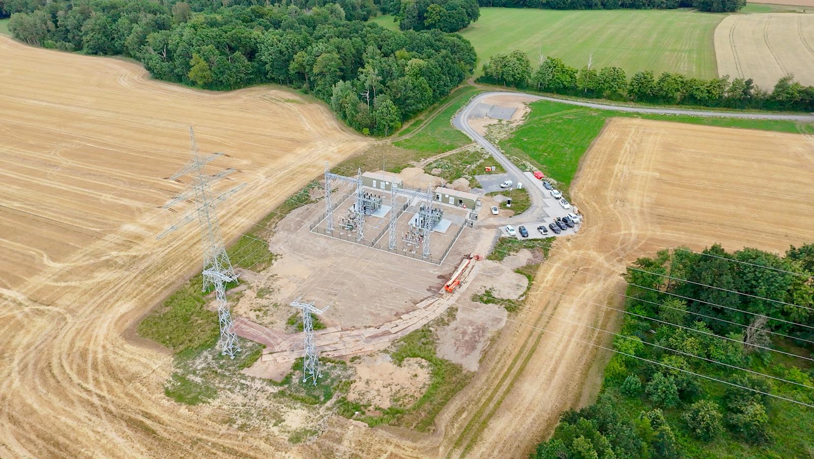 Luftaufnahme eines Umspannwerks mit Hochspannungsanlagen, Portalen und Netzanschlusspunkt zur Anbindung an das bestehende Stromnetz. Das Umspannwerk liegt zwischen landwirtschaftlichen Flächen und ist über eine Zufahrtsstraße erreichbar.