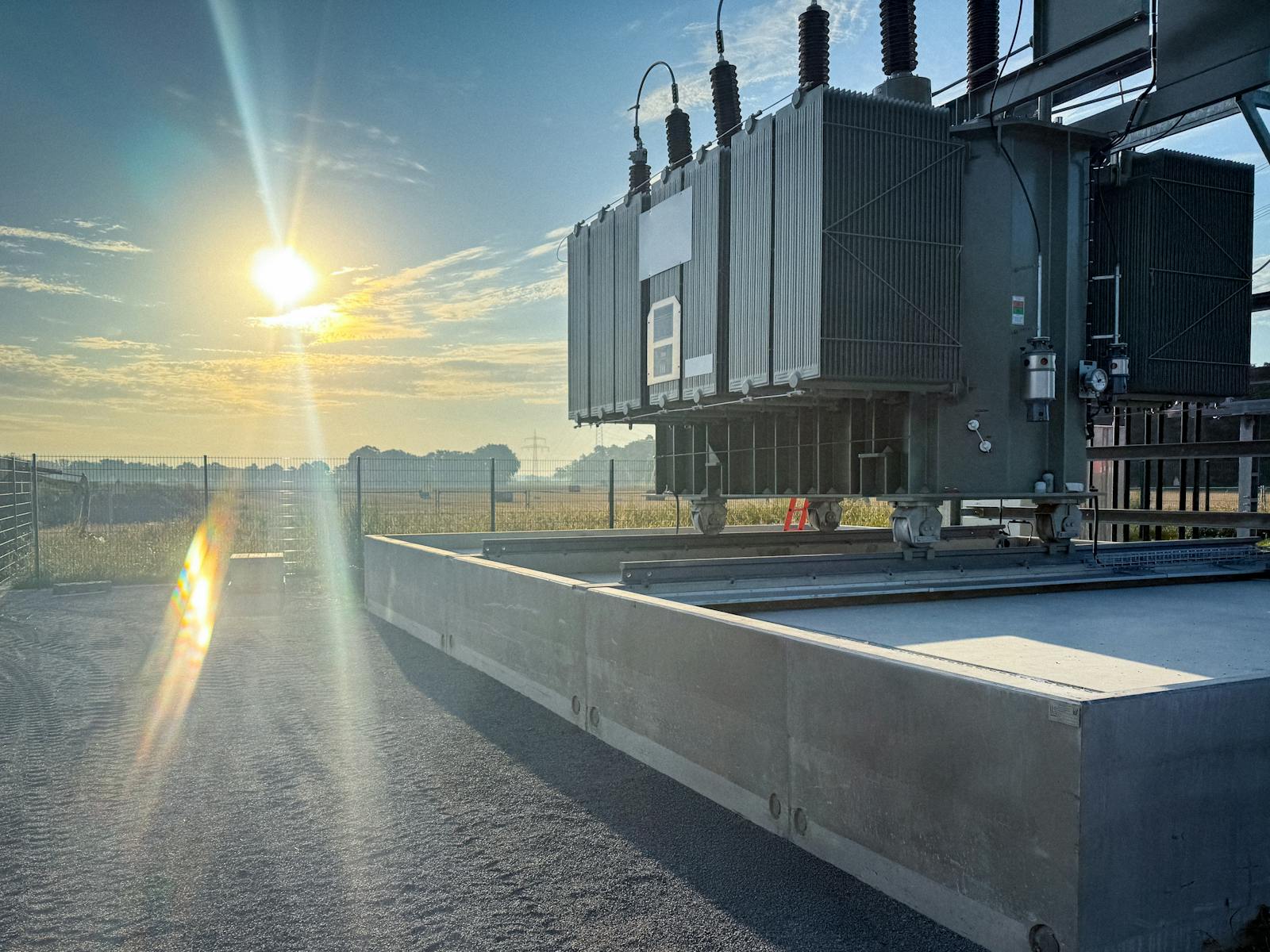 Seitenansicht eines Leistungstransformators in einem Umspannwerk bei tief stehender Sonne. Der Transformator mit Kühlradiatoren und Hochspannungsdurchführungen steht auf einem Betonfundament mit Ölauffangwanne.