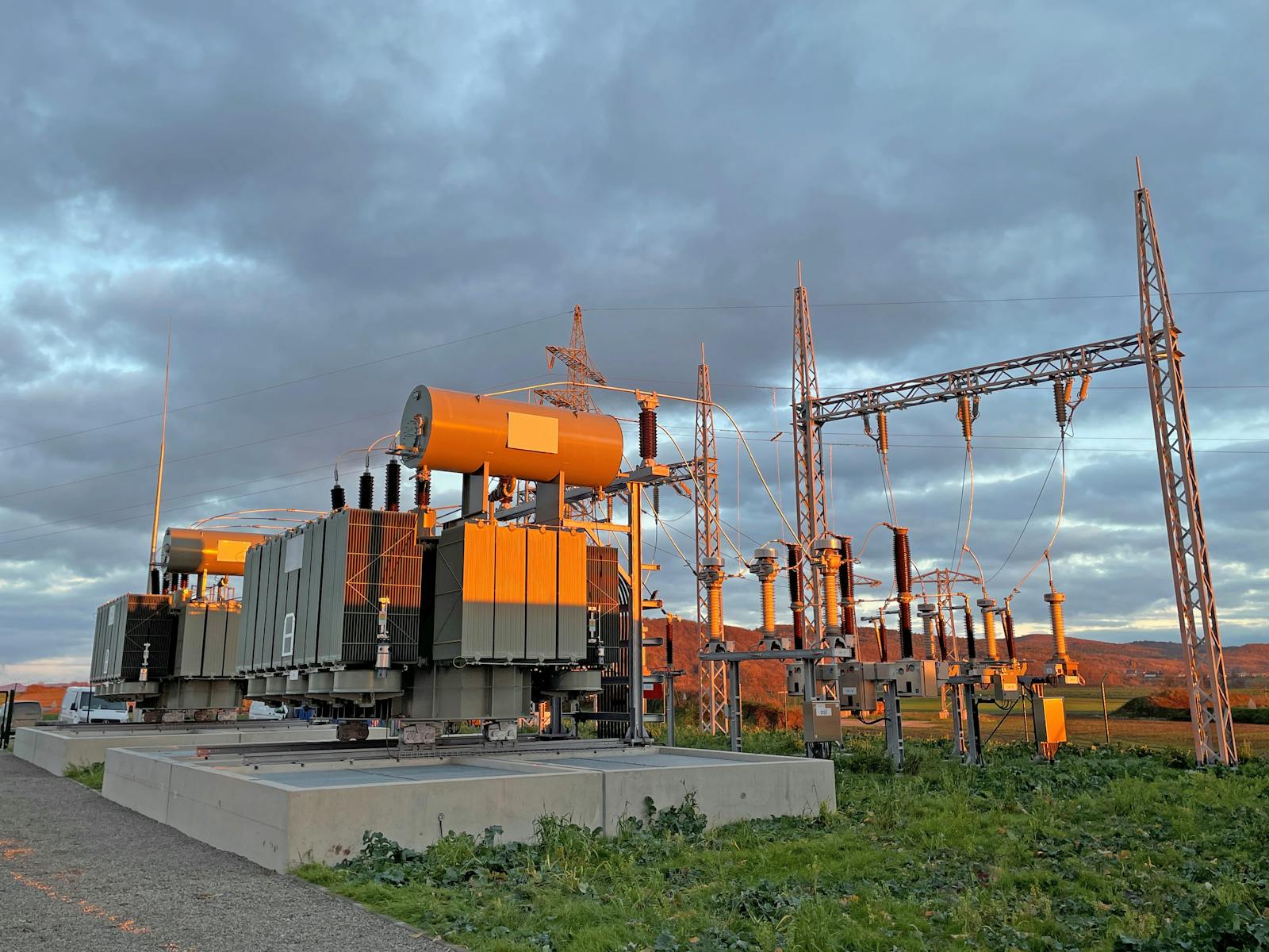 Zwei Leistungstransformatoren mit Kühlradiatoren und Ausdehnungsgefäßen in einem Umspannwerk bei tief stehender Abendsonne. Rechts sind Hochspannungsschaltanlagen, Isolatoren und ein Leitungsportal zur Freileitungsanbindung zu sehen.