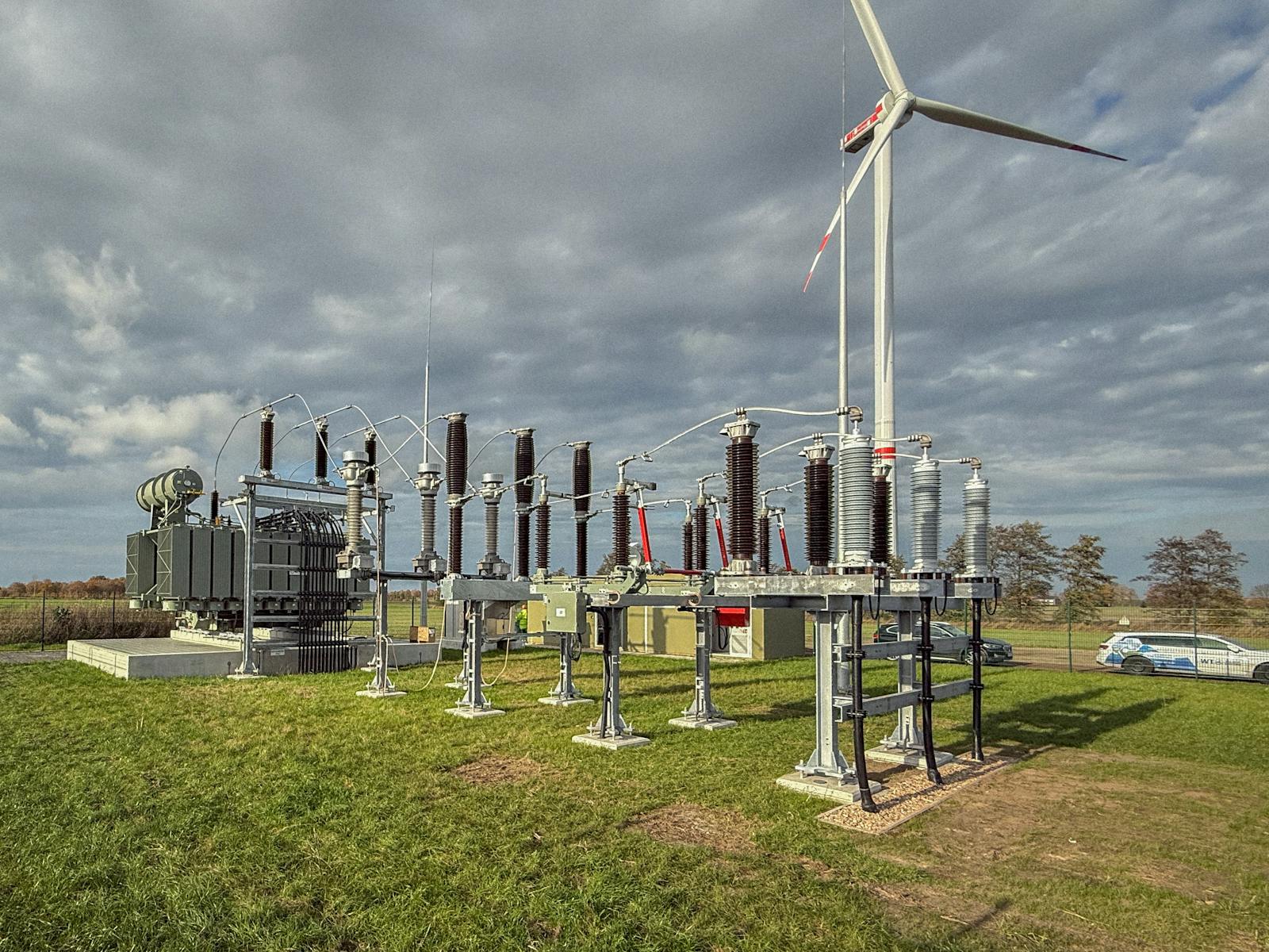Umspannwerk zur Netzanbindung einer Windenergieanlage mit Leistungstransformator, Hochspannungsschaltanlagen und Isolatoren. Im Hintergrund ist eine Windkraftanlage unter bewölktem Himmel zu sehen.