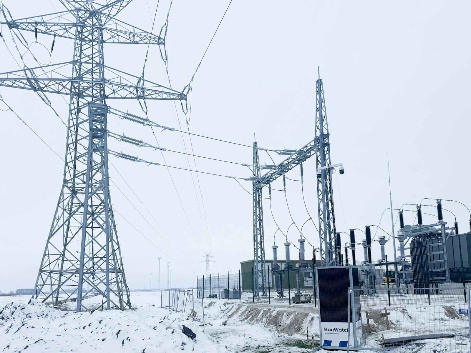 Verschneites Umspannwerk mit Leistungstransformator, Hochspannungsschaltanlagen und Stahlgitterportal zur Anbindung an das Stromnetz über eine Hochspannungsfreileitung.