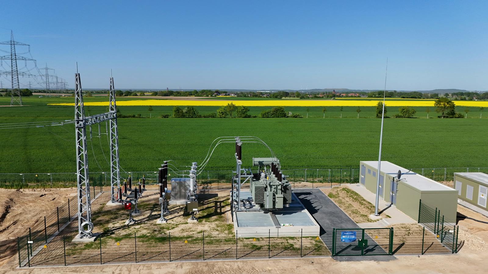 Luftaufnahme eines Umspannwerks mit Leistungstransformator, Hochspannungsschaltanlagen, Stahlgitterportal zur Freileitungsanbindung und Betriebsgebäuden, umgeben von landwirtschaftlichen Flächen.