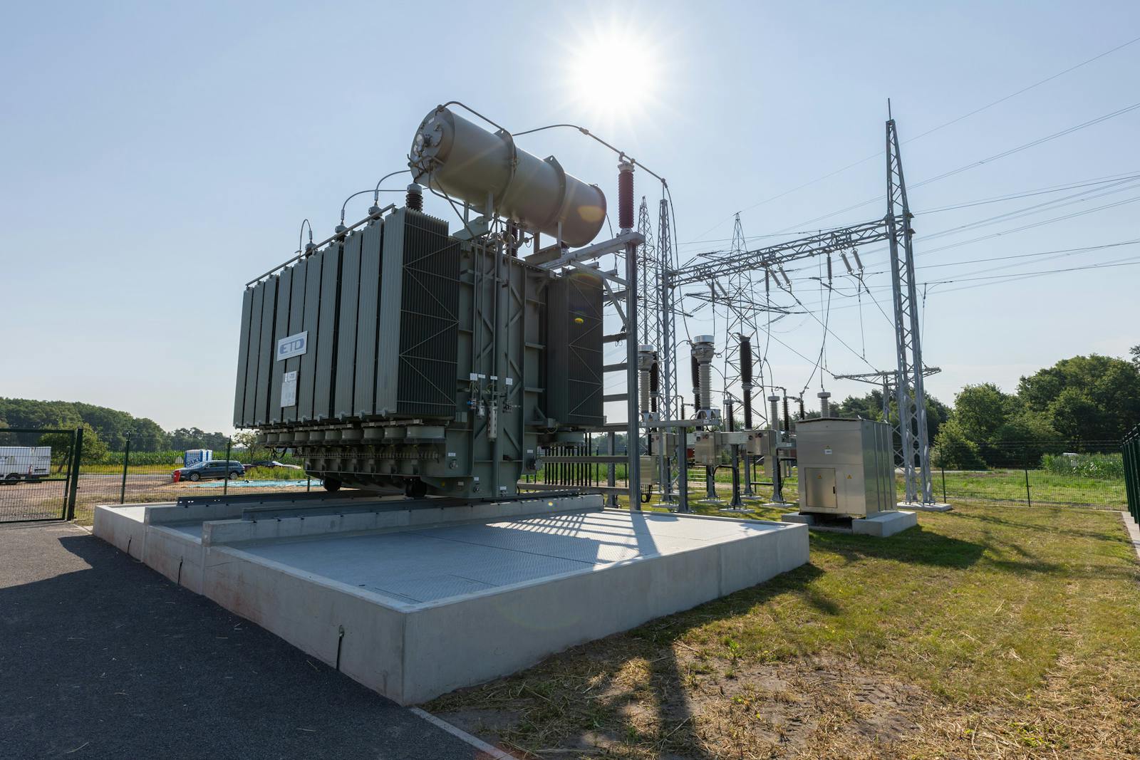 Power transformer with cooling radiators, high-voltage bushings and an oil conservator tank at an electrical substation. The transformer is installed on a concrete foundation with an oil containment basin. High-voltage switchgear, a single overhead line gantry and overhead power lines are visible in the background.