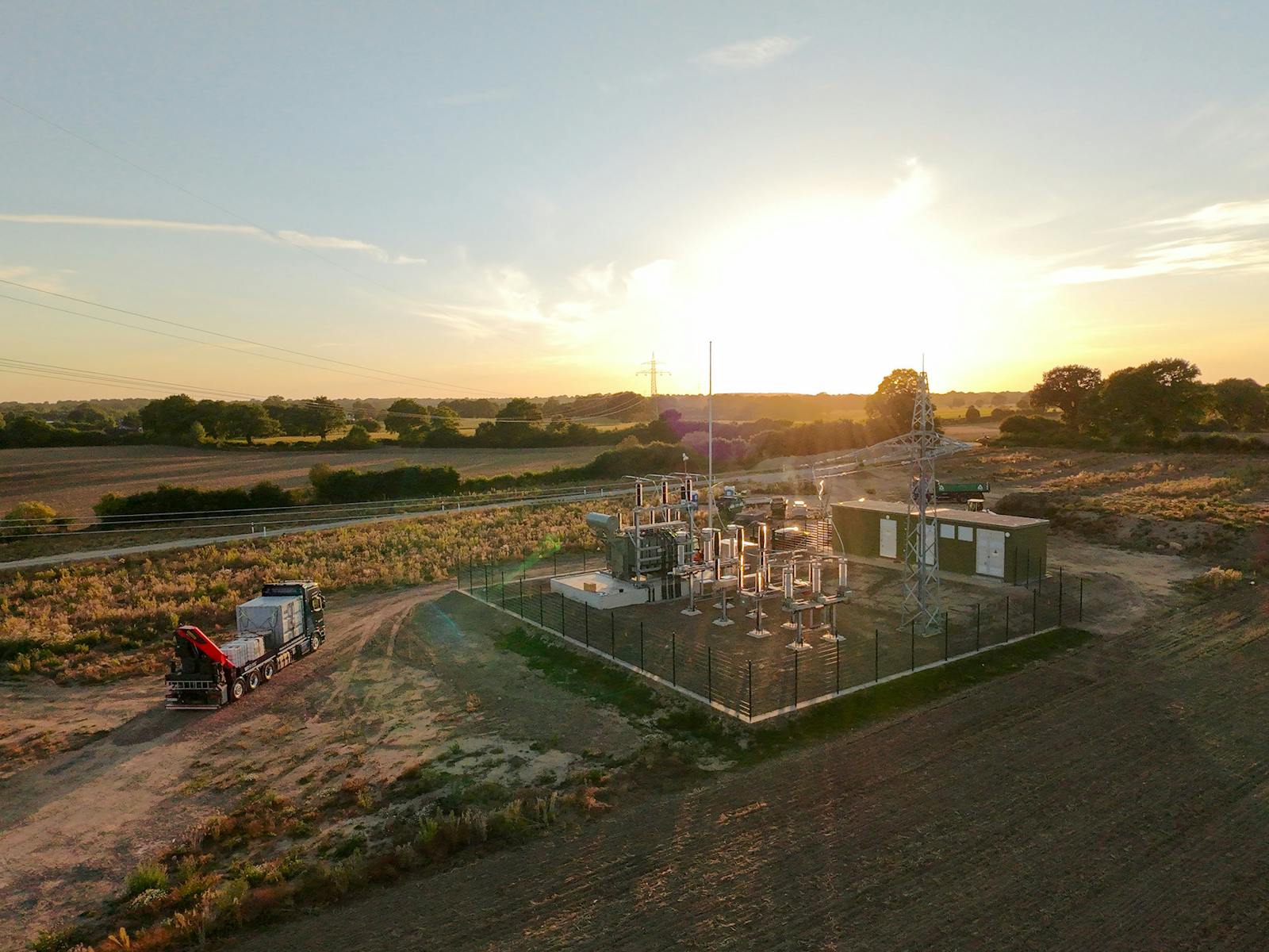 Luftaufnahme eines Umspannwerks bei Sonnenuntergang mit Leistungstransformator, Hochspannungsschaltanlage, Leitungsportal und Betriebsgebäude. Die Anlage liegt inmitten einer ländlichen Landschaft und ist über Freileitungen an das Stromnetz angebunden.