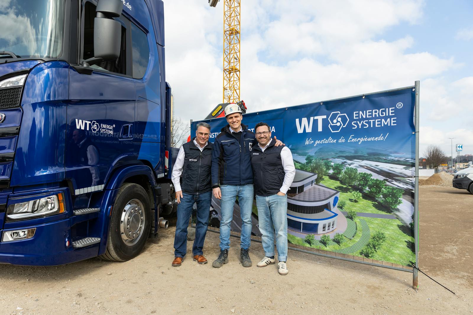 Gruppenfoto von drei Personen vor einem Lkw und einem Baustellenbanner von WT Energiesysteme mit einer Visualisierung des neuen Bürokomplexes am Standort Riesa.