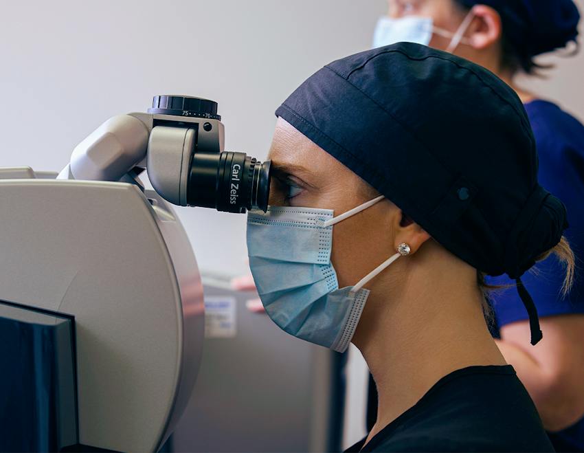 Two masked clinicians operating ophthalmic diagnostic equipment during an eye procedure.