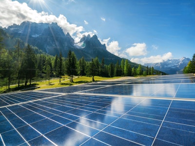 Solar panels in a field with pine trees and mountains under a blue sky.