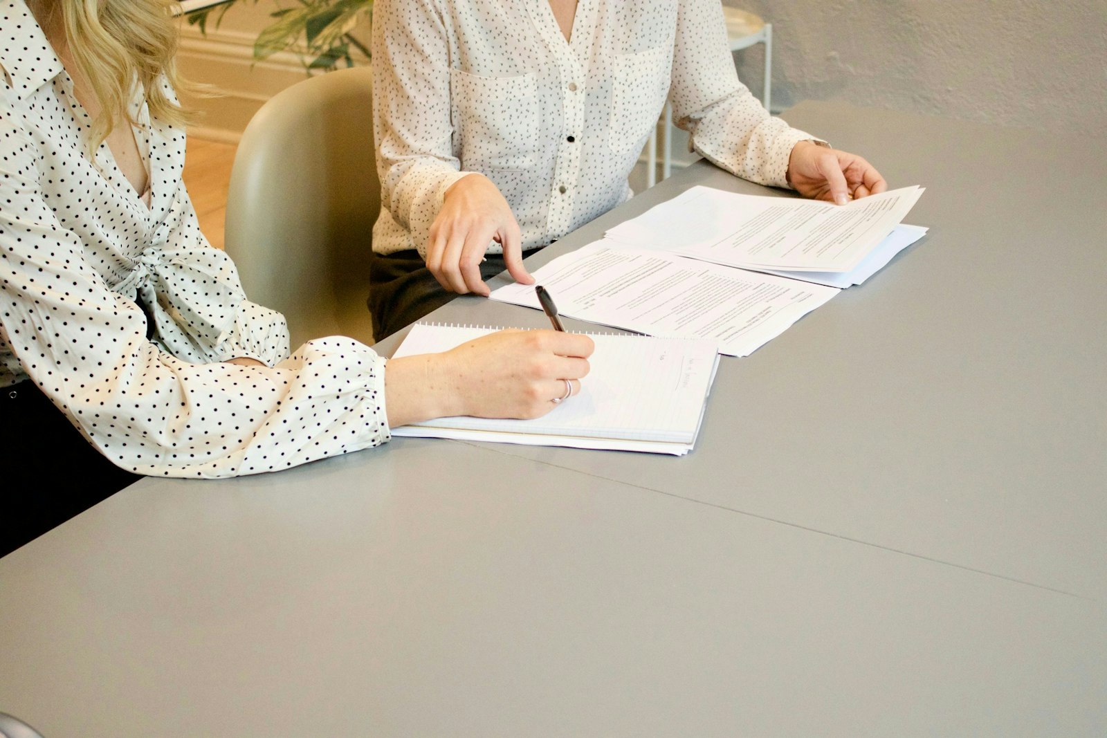 Woman signing on white printer paper beside woman about to touch the documents.