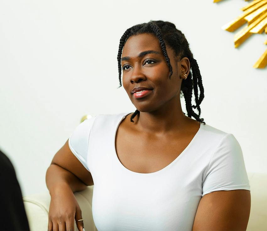 Woman with braided hair sitting relaxed in white top, smiling softly.