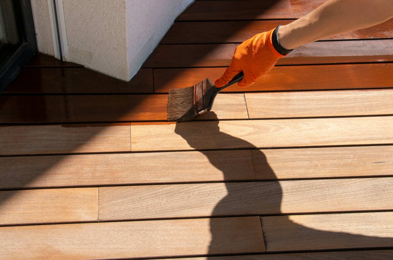 A person wearing an orange glove brushes a brown stain onto planks of wood.