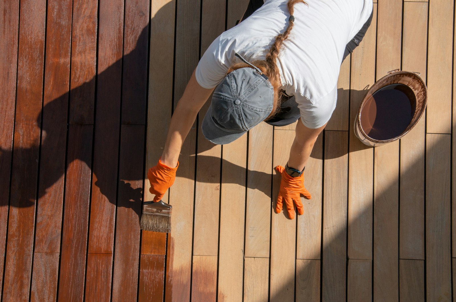 An overhead shot of a worker in a white shirt and grey hat panting a brown stain on a deck.