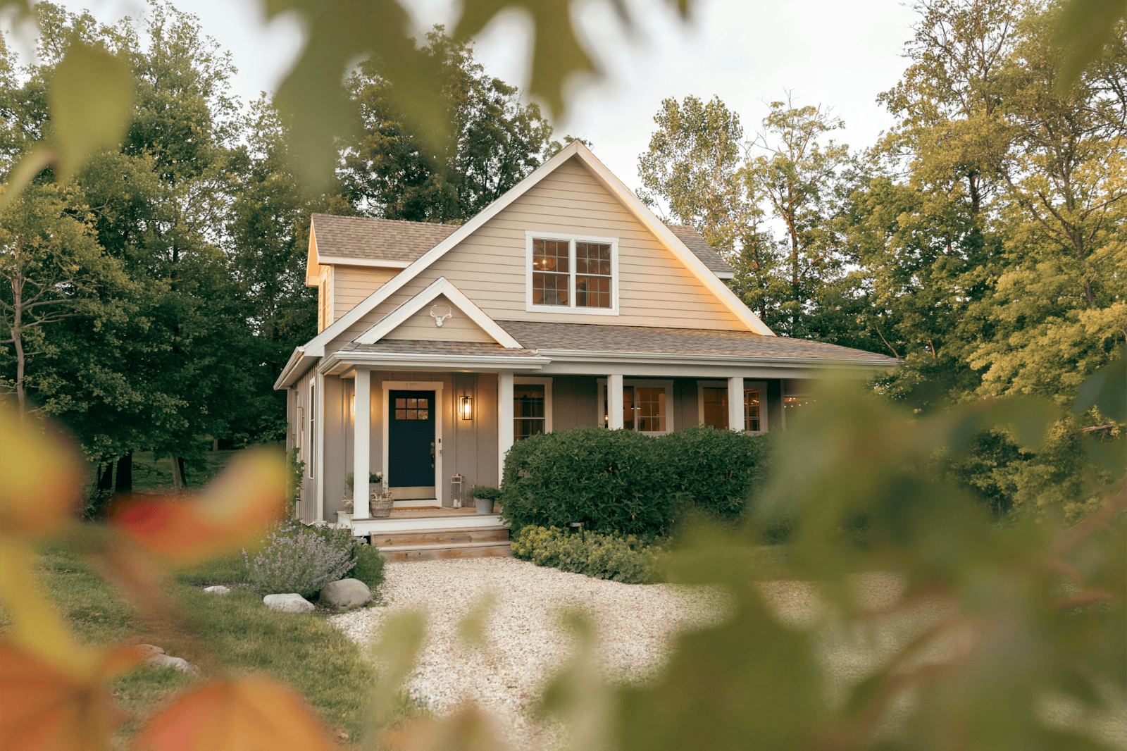 A beige house sits beyond a gravel lot on a treed lot.