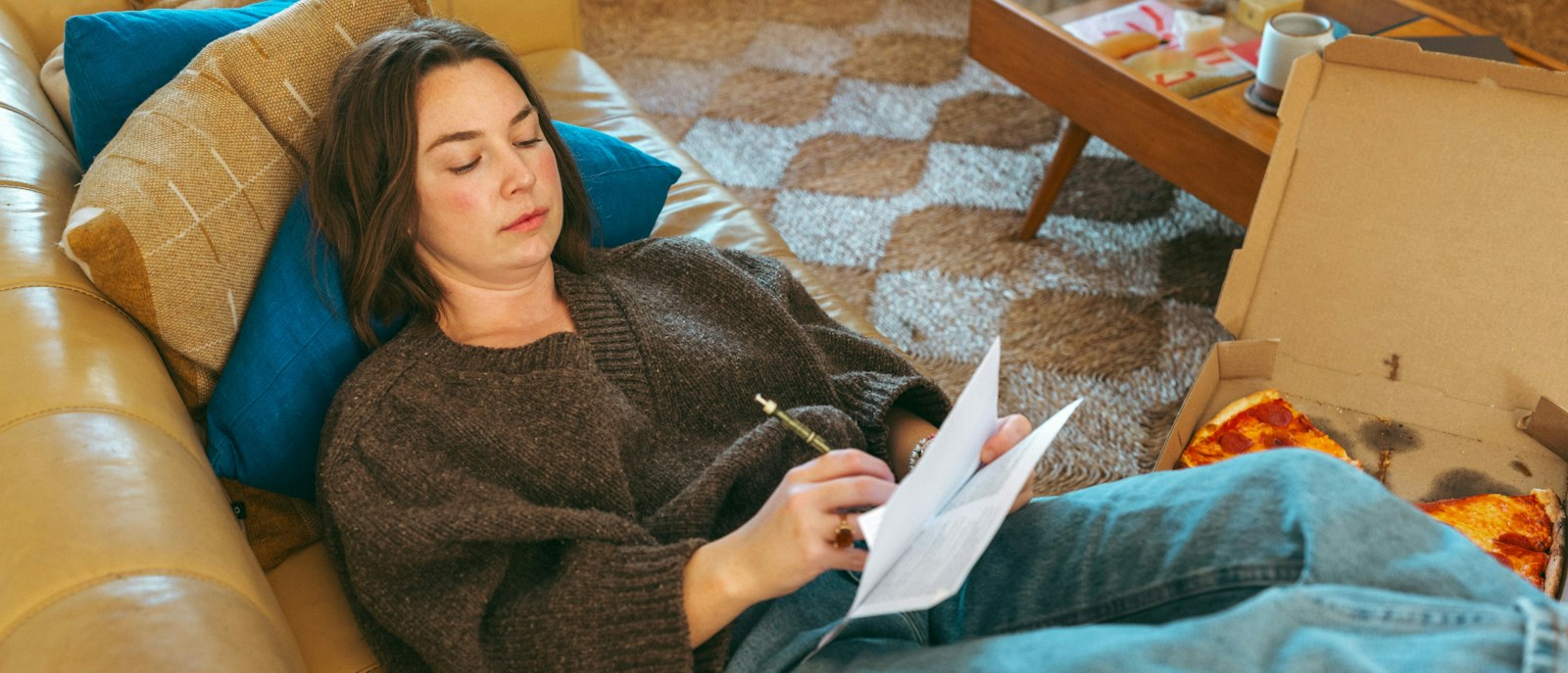 A woman wearing a brown sweater looks at a piece of paper while lying on a couch.