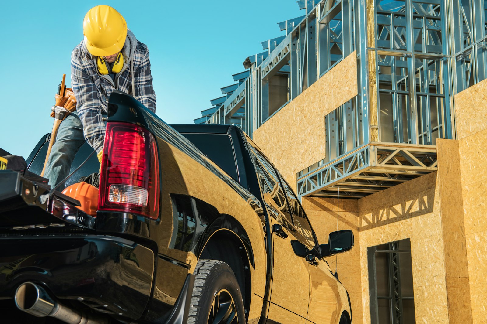A contractor wearing a yellow safety helmet stands in the bed of a black pickup truck in front of a construction site.