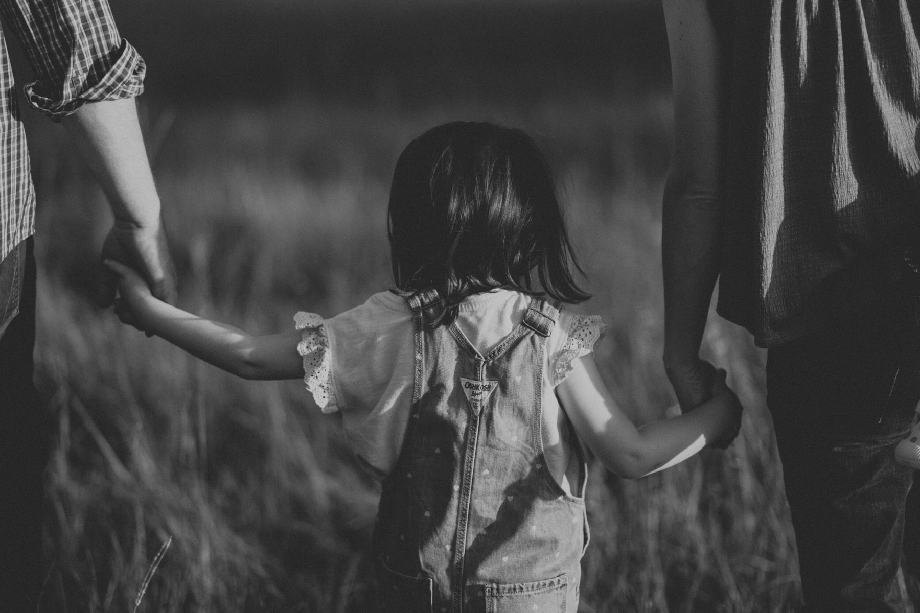 Black and white photo of a toddler holding both parents' hands during a summer family session in a meadow