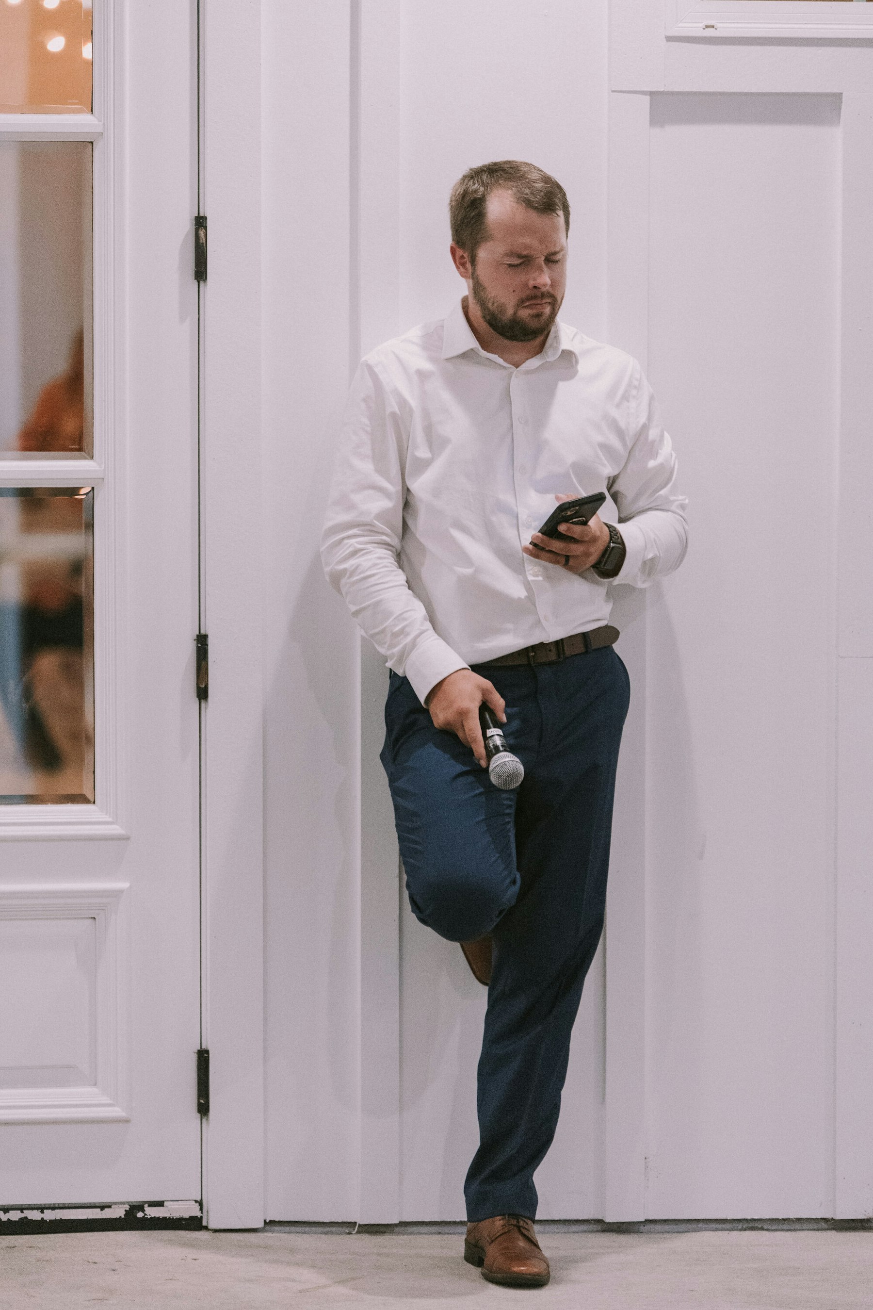 Groom leaning in a doorway holding a microphone at a white barn wedding