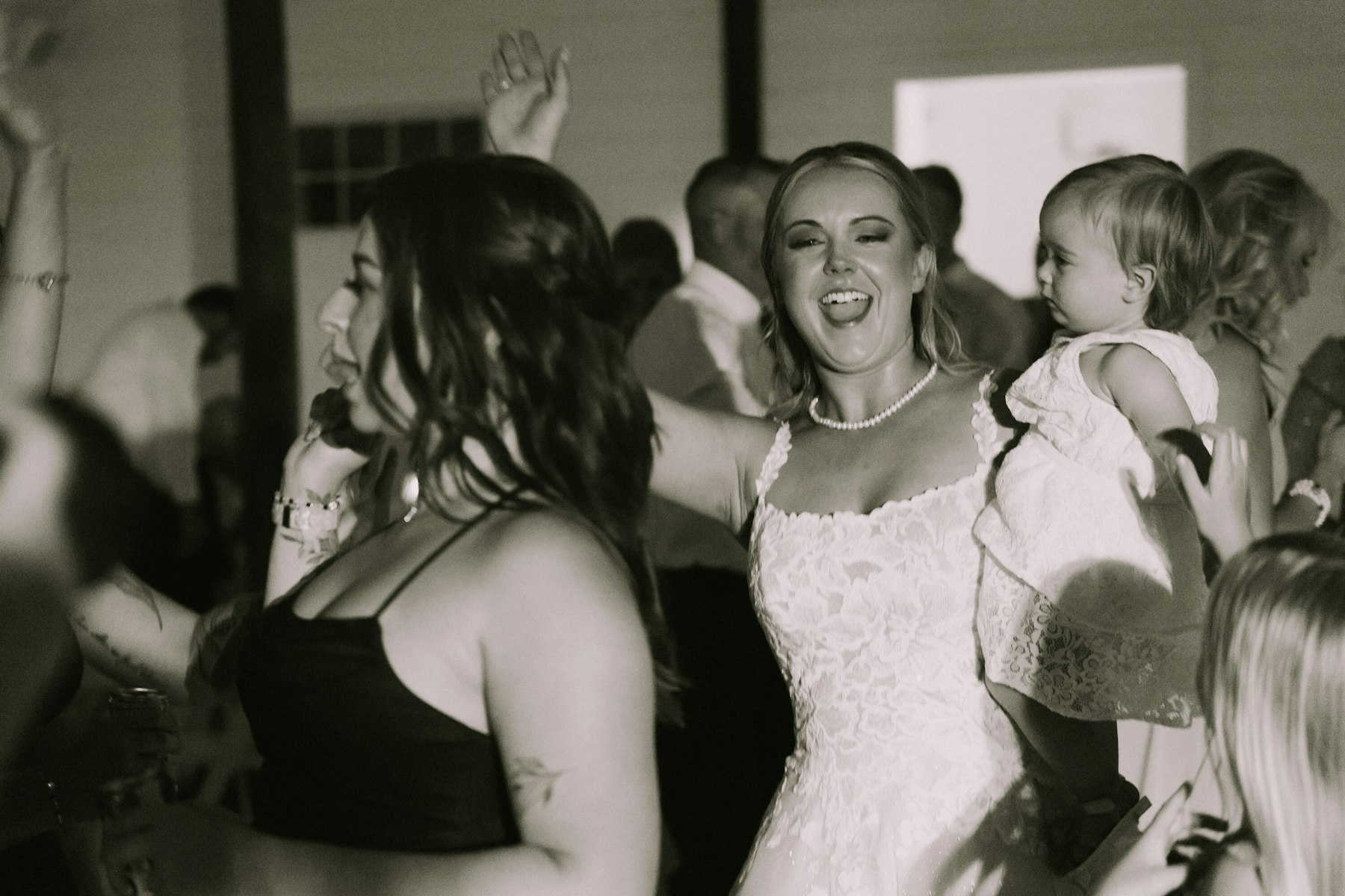Black and white photo of the bride dancing with guests and holding a baby at a white barn wedding