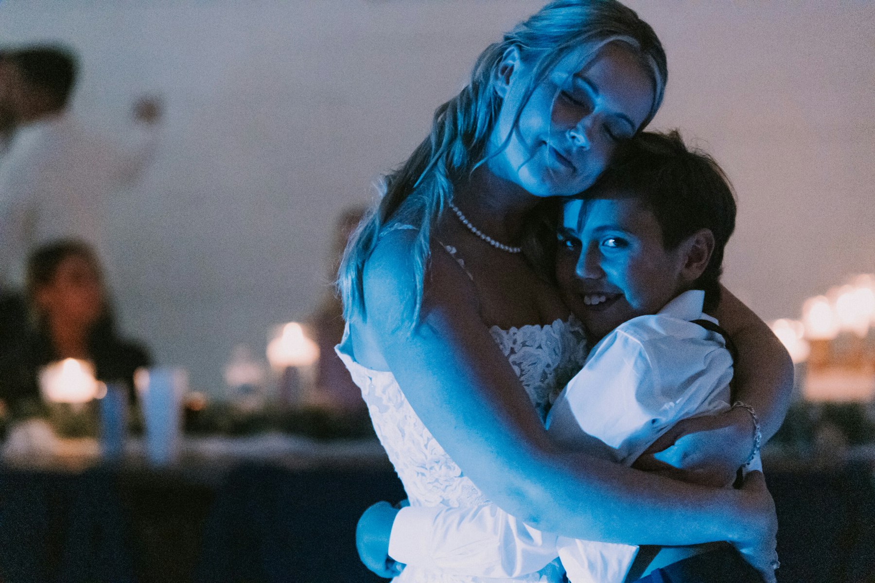 Bride hugging a young guest in blue reception light at a white barn wedding
