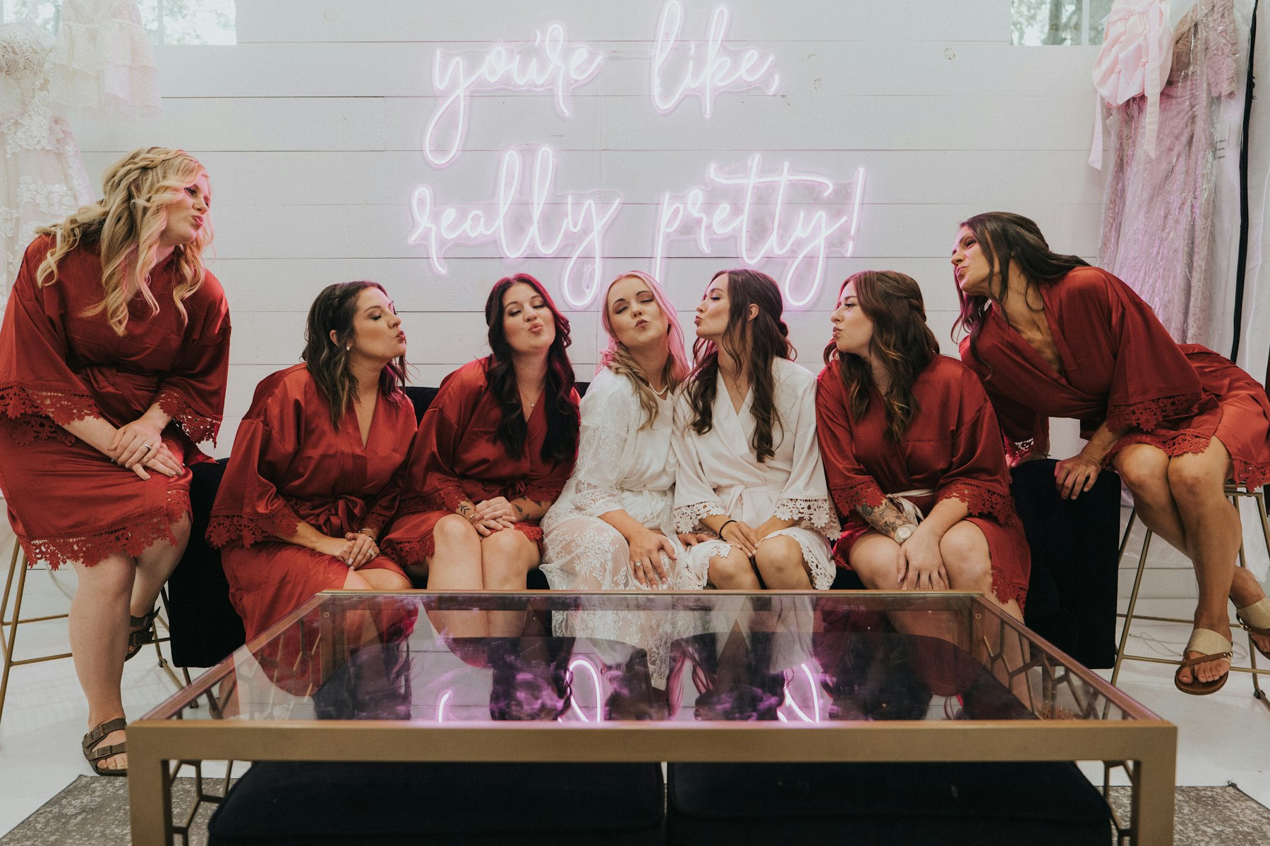 Bridesmaids in burgundy robes under a neon sign at a white barn wedding