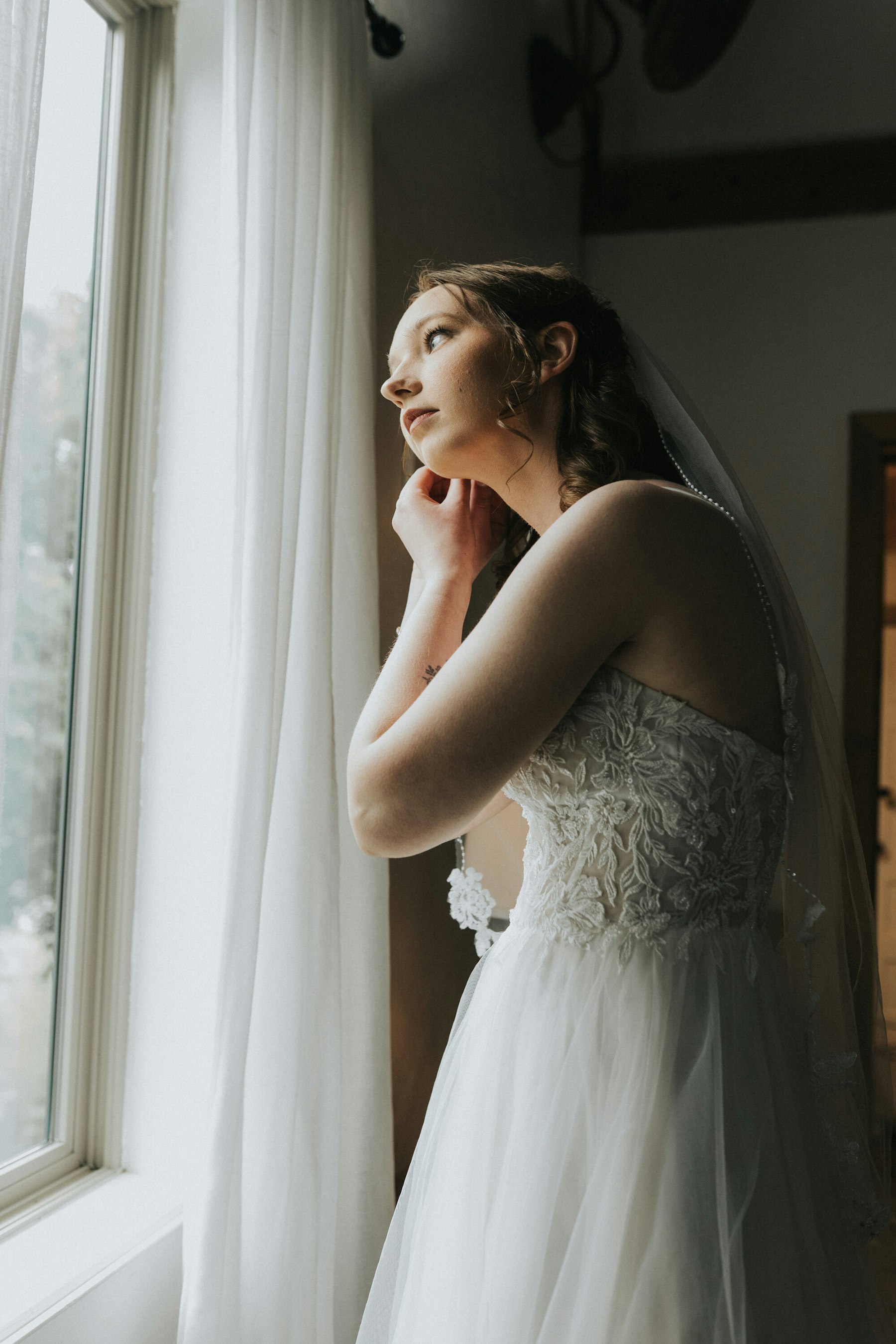 Bride putting on her earrings at the window at a rustic barn wedding
