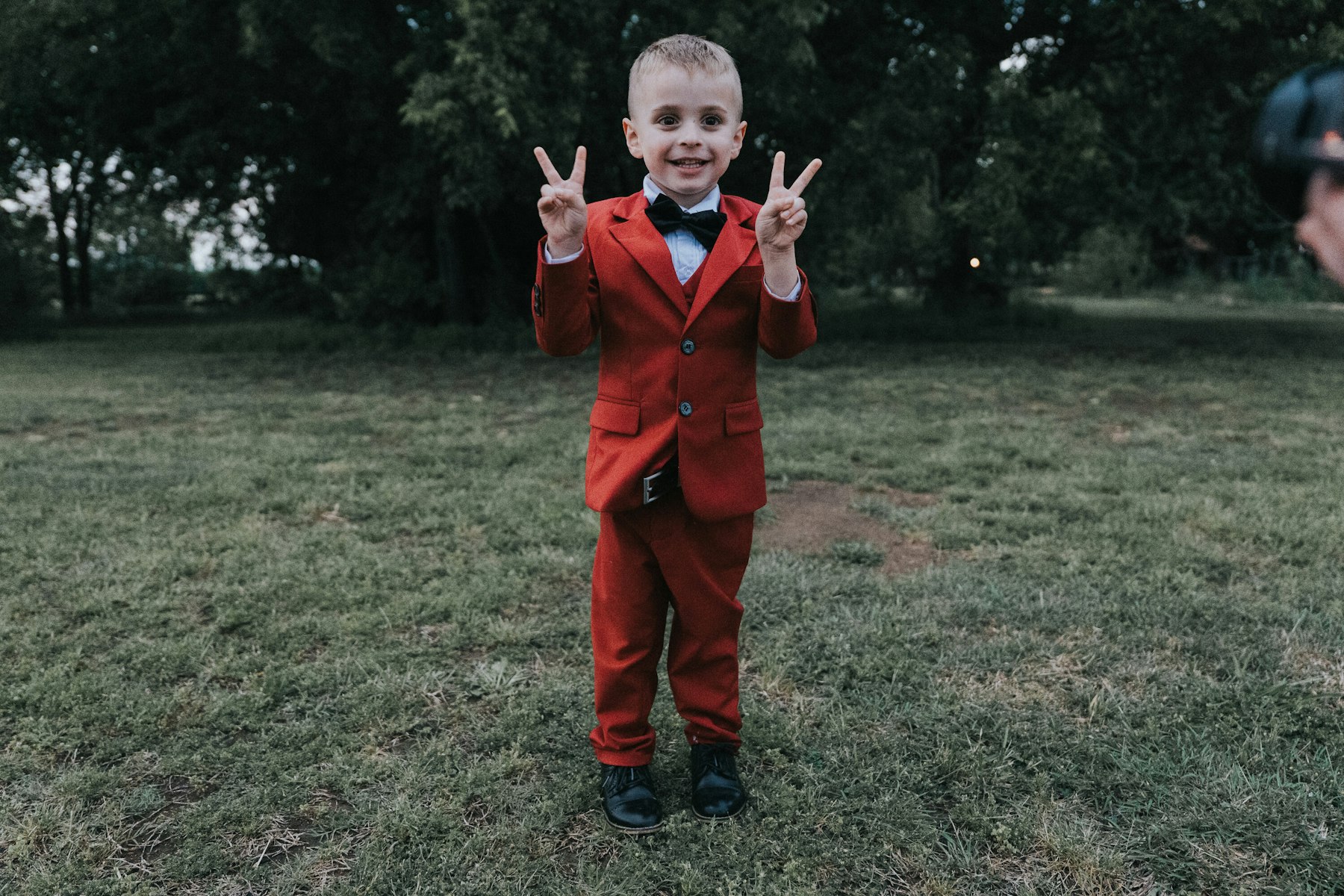 Ring bearer in a red suit flashing peace signs at a rustic barn wedding