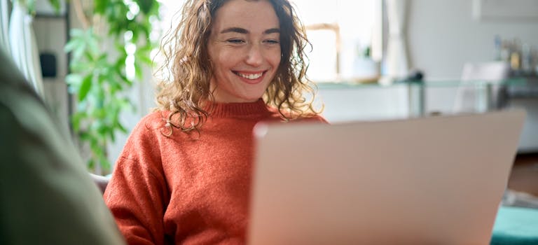 Woman in orange jumper smiling while looking at laptop in her apartment